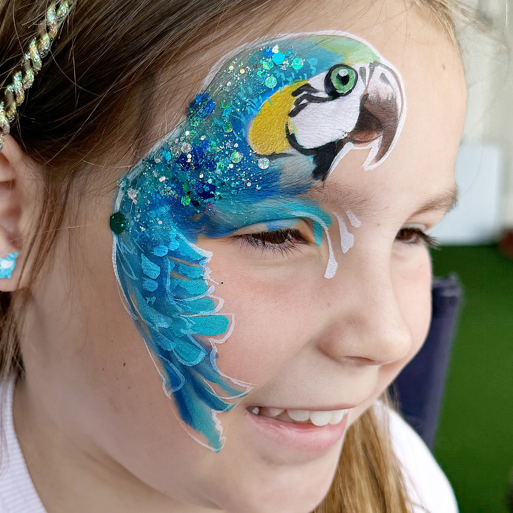 Dziewczynka z pomalowaną twarzą w motyw błękitnej papugi z brokatem, widok z profilu. Kreatywny makijaż artystyczny na imprezę dla dzieci.