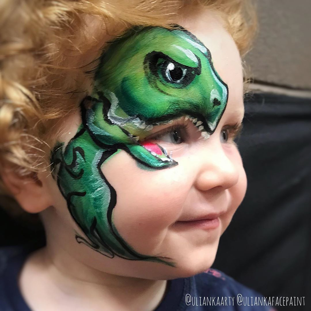 Chłopiec z pomalowaną twarzą w zielonego dinozaura, widok z profilu, artystyczny makijaż na skórze, kreatywna stylizacja, jasne włosy, uśmiech.