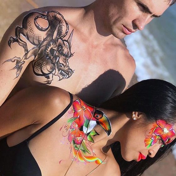 Para z artystycznym body paintingiem: mężczyzna z motywem smoka, kobieta z tukanem i kwiatami. Plaża w tle, jasne światło.