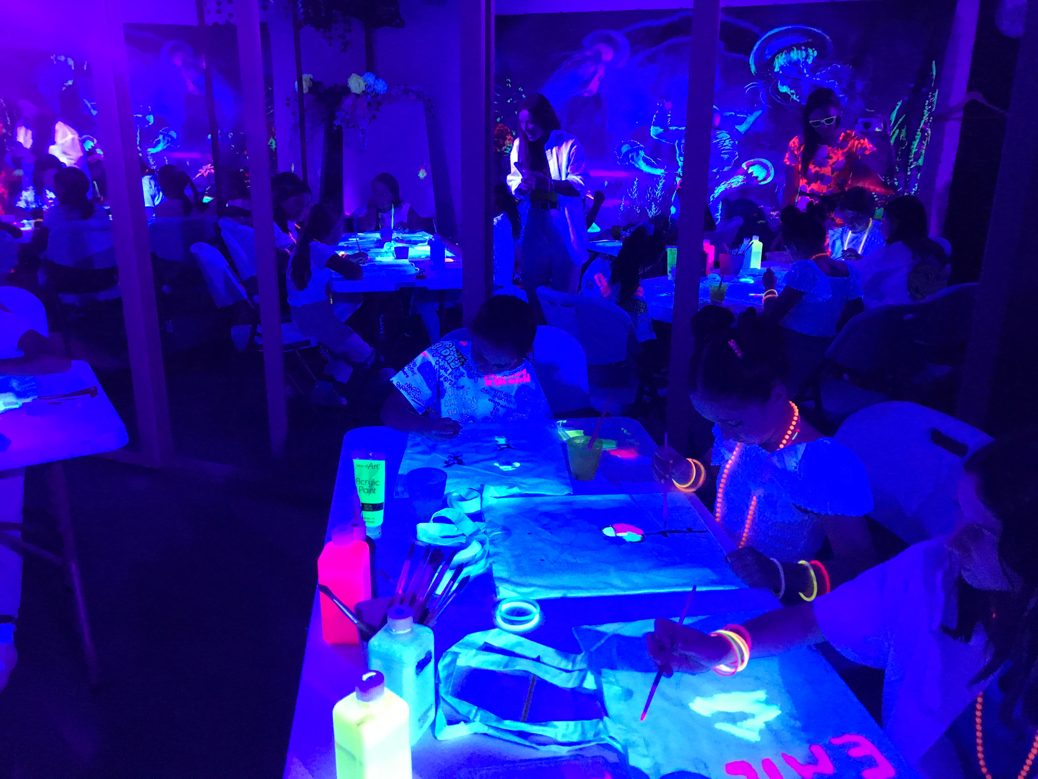 Uczestnicy warsztatów malarskich UV tworzą obrazy w ciemnym pomieszczeniu oświetlonym światłem UV. Widoczne farby neonowe, pędzle i płótna.