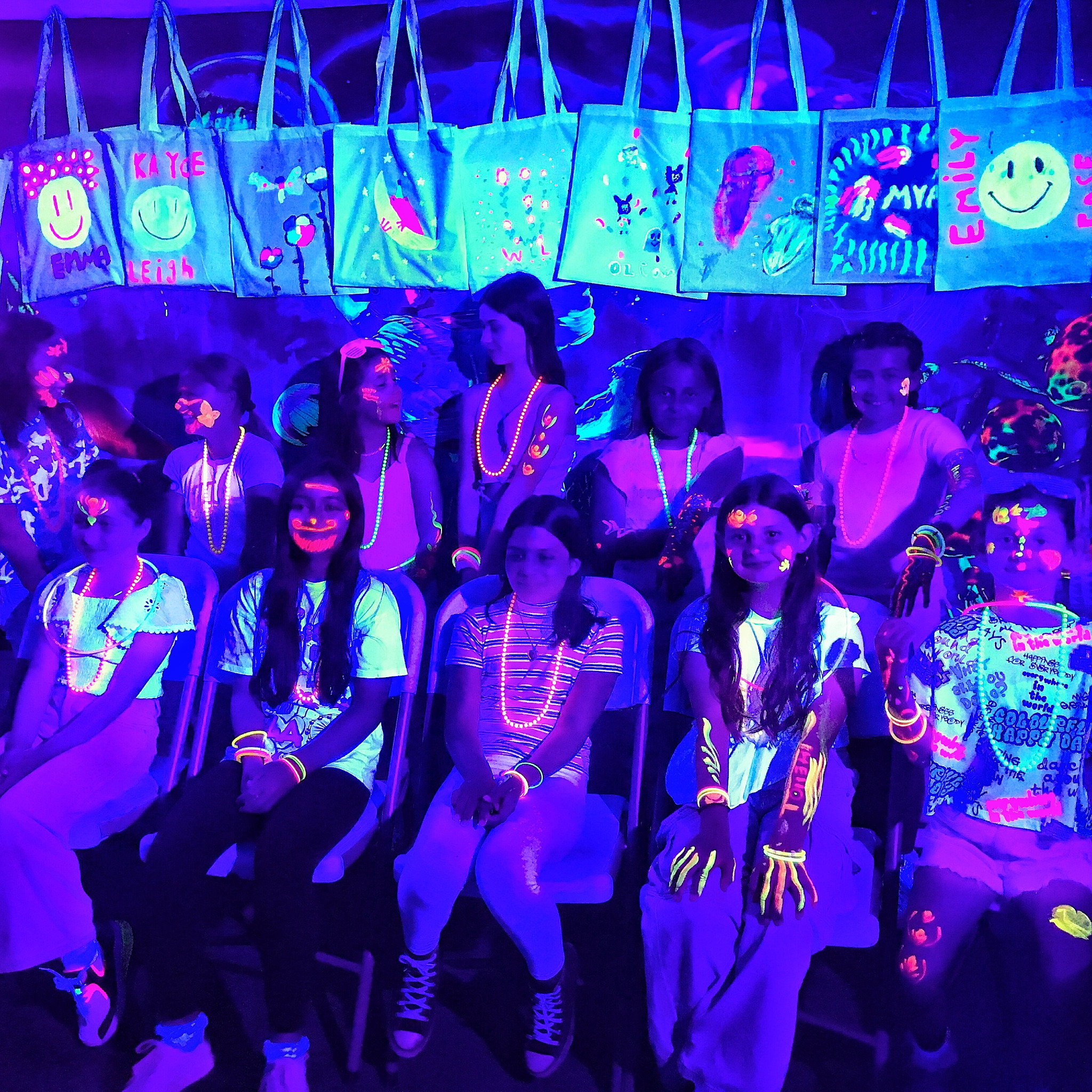 Grupa dzieci w świetle UV, siedzących na krzesłach, z pomalowanymi twarzami i torbami z własnoręcznymi rysunkami, zawieszonymi w tle. Kreatywne warsztaty.