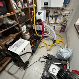 HydroTech Południe osuszanie po zalaniu, wynajem osuszaczy, likwidacja szkód po awari, lokalizacja wycieków - Osuszacz kondensacyjny w pomieszczeniu z instalacją grzewczą i regałem. Żółte węże, sprzęt do ćwiczeń. Sprzęt do osuszania po zalaniach w trakcie pracy.