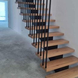 Iwona Cierpioł - Nowoczesne schody z drewnianymi stopniami i czarną, metalową balustradą. Minimalistyczny design, surowy beton na podłodze, widok z dołu na górę.