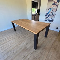 Iwona Cierpioł - Stół z blatem z jasnego drewna i czarnymi, ukośnymi nogami, ustawiony w nowoczesnym wnętrzu z drewnopodobną podłogą i dekoracyjnym obrazem na ścianie.