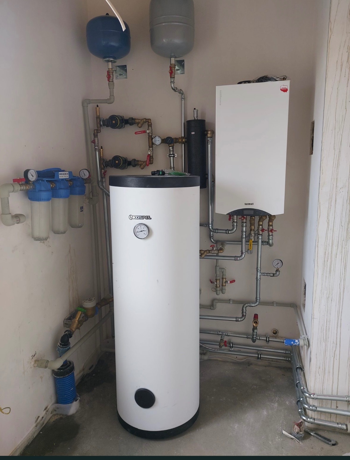 Biały bojler Kospel z podłączeniami, filtry wody, zbiorniki wyrównawcze i piec Termet na ścianie. Instalacja hydrauliczna w kotłowni, widok z dołu.
