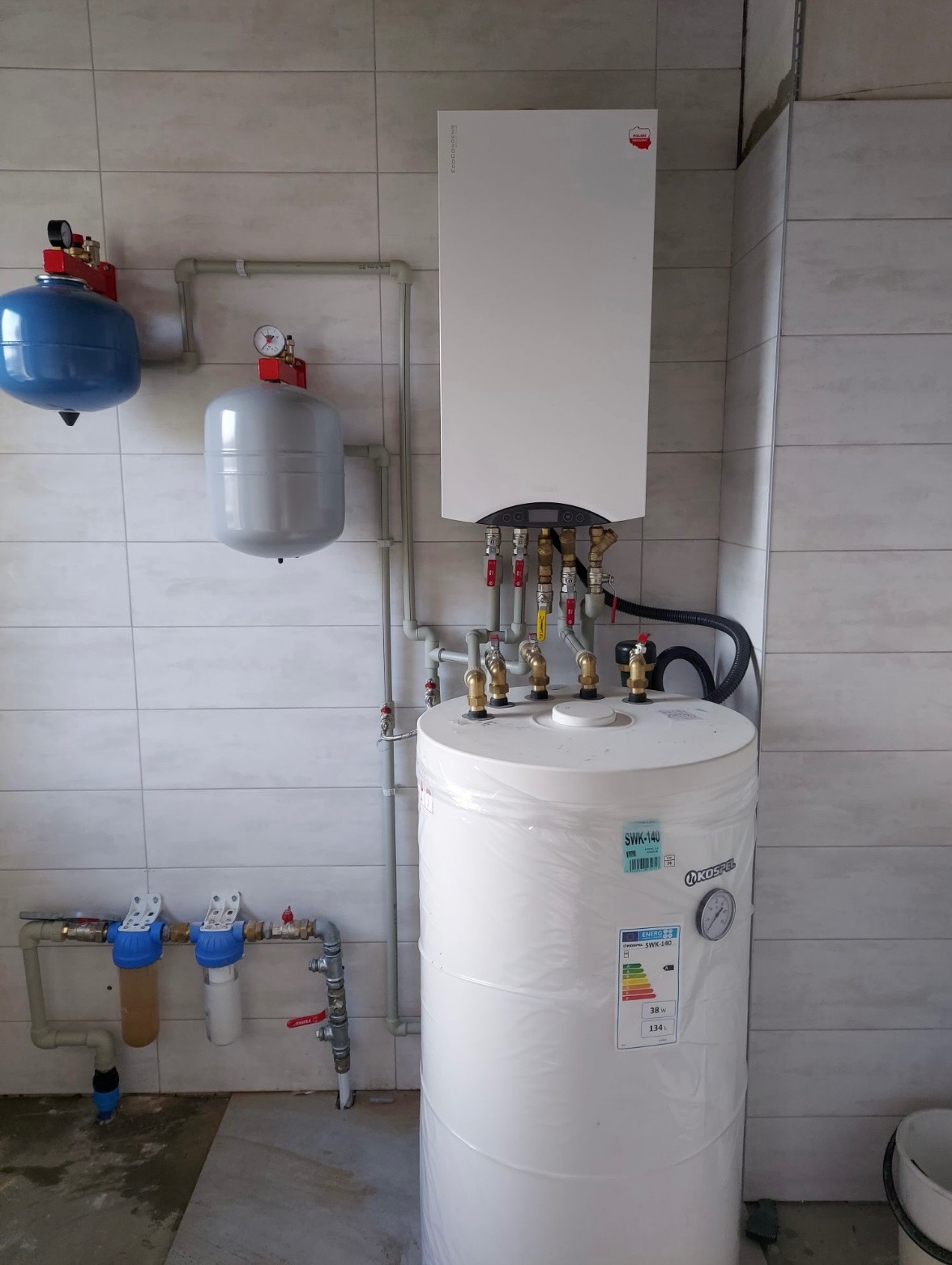 Biały bojler Kospel SWK-140 z podłączonymi rurami i zaworami, zamontowany w pomieszczeniu z jasnymi płytkami. Widoczne filtry wody i zbiorniki wyrównawcze.