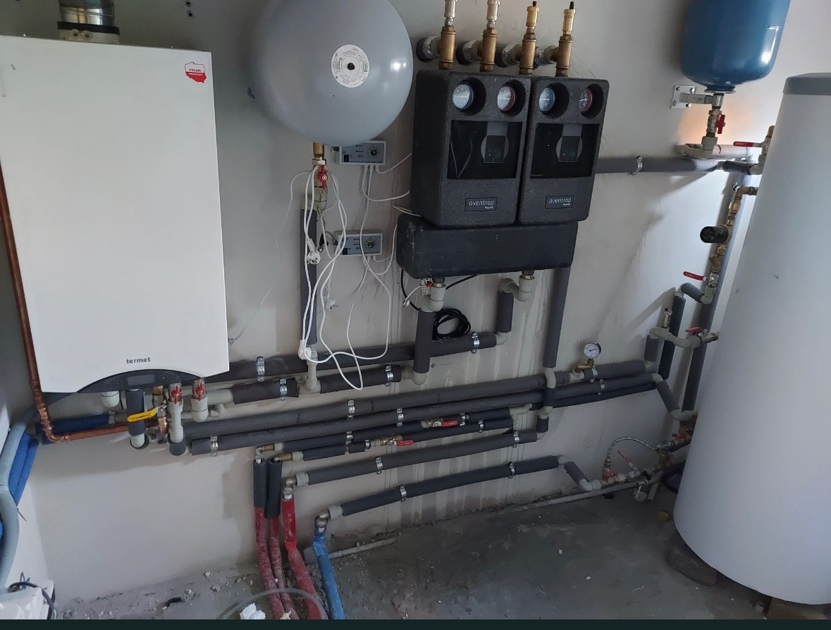 Złożona instalacja grzewcza z kotłem Termet, zbiornikiem wyrównawczym i rozdzielaczem Aventrop. Rury izolowane, liczne zawory i manometry. Nowoczesny system w kotłowni.