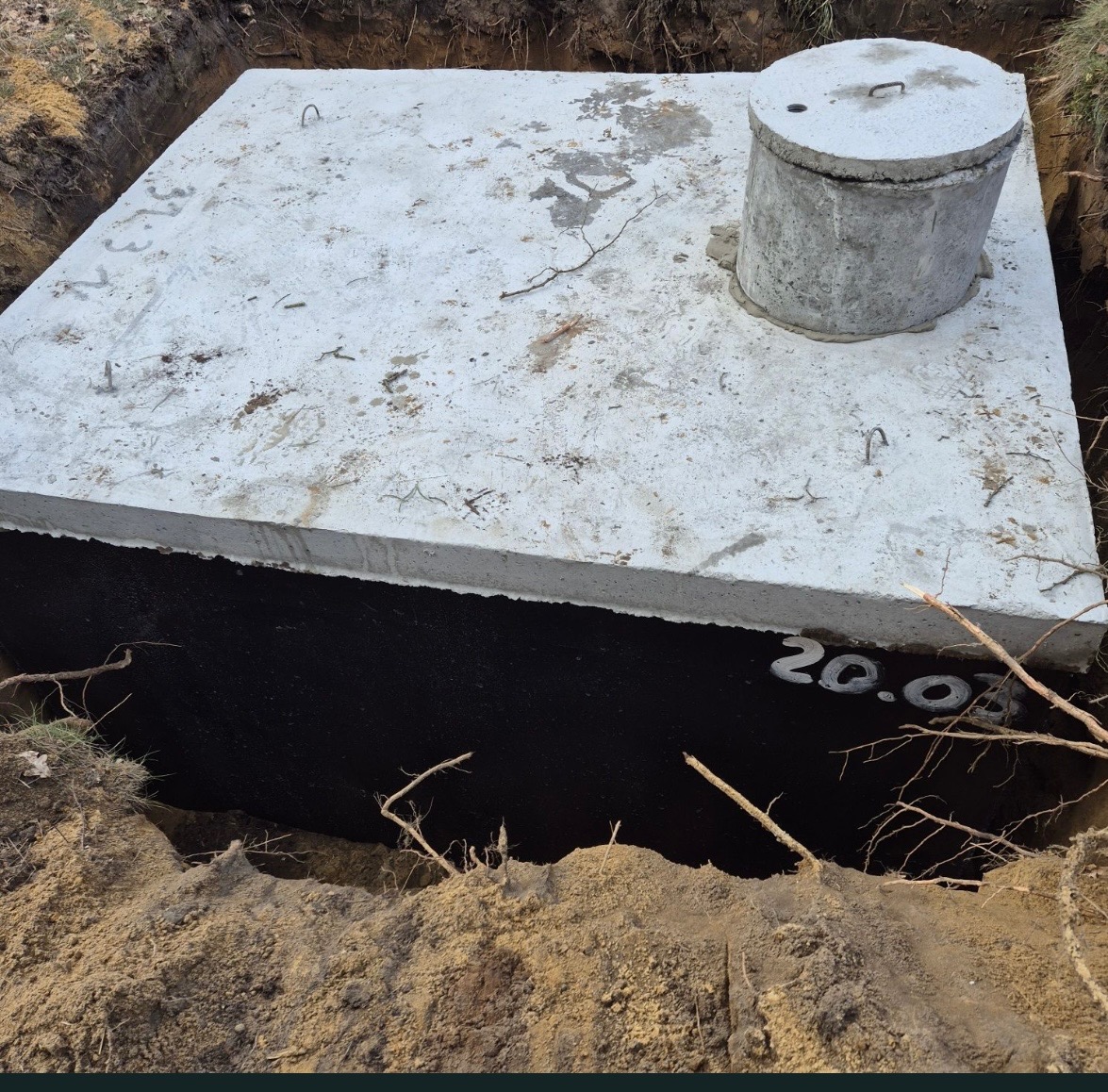 Betonowy zbiornik szamba z pokrywą, częściowo zakopany w ziemi, z widoczną izolacją bitumiczną i napisem '20.02' na boku, otoczony wykopem z ziemią.