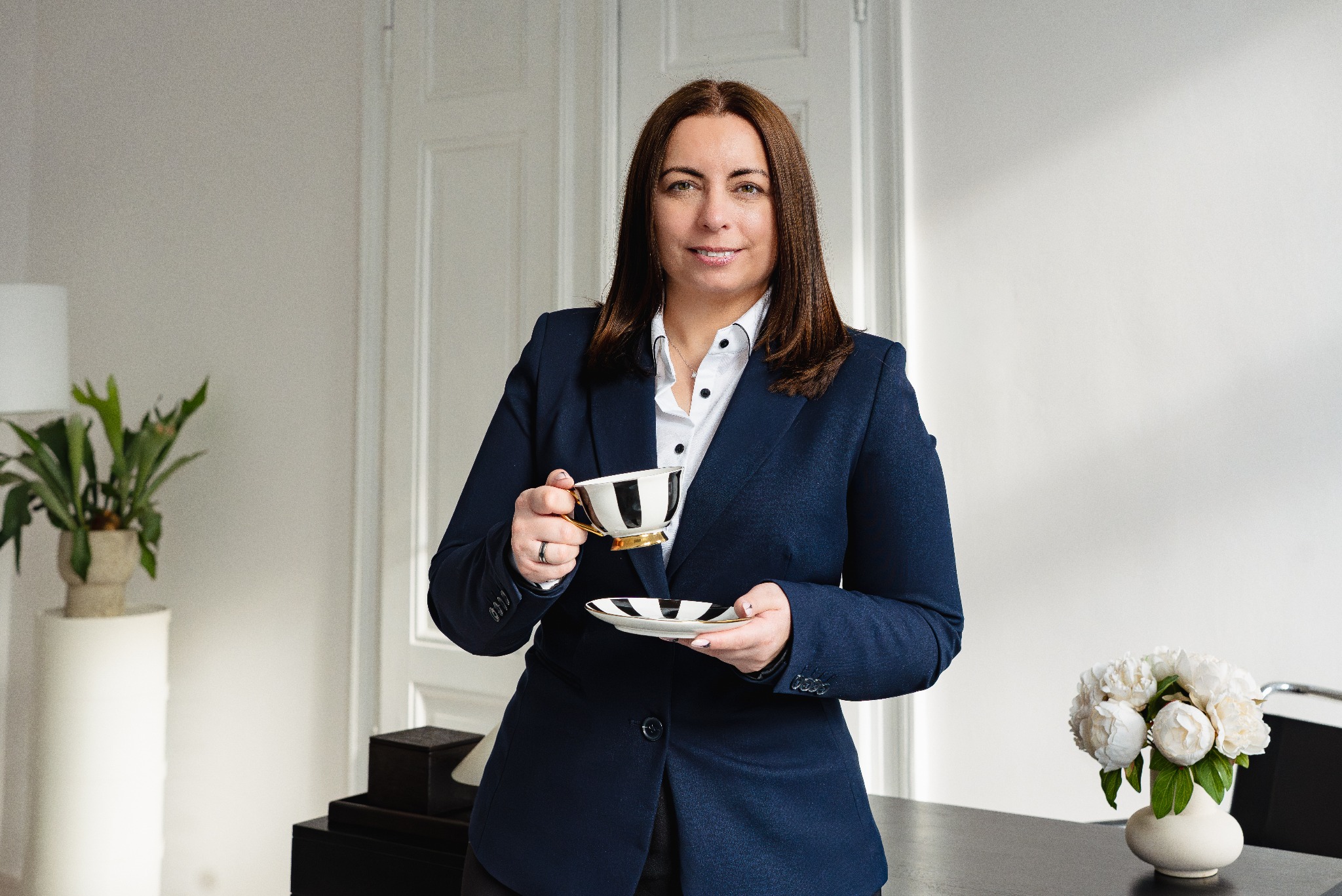 Profesjonalistka w granatowej marynarce pije kawę z eleganckiej filiżanki w biurze. W tle białe drzwi i rośliny doniczkowe.