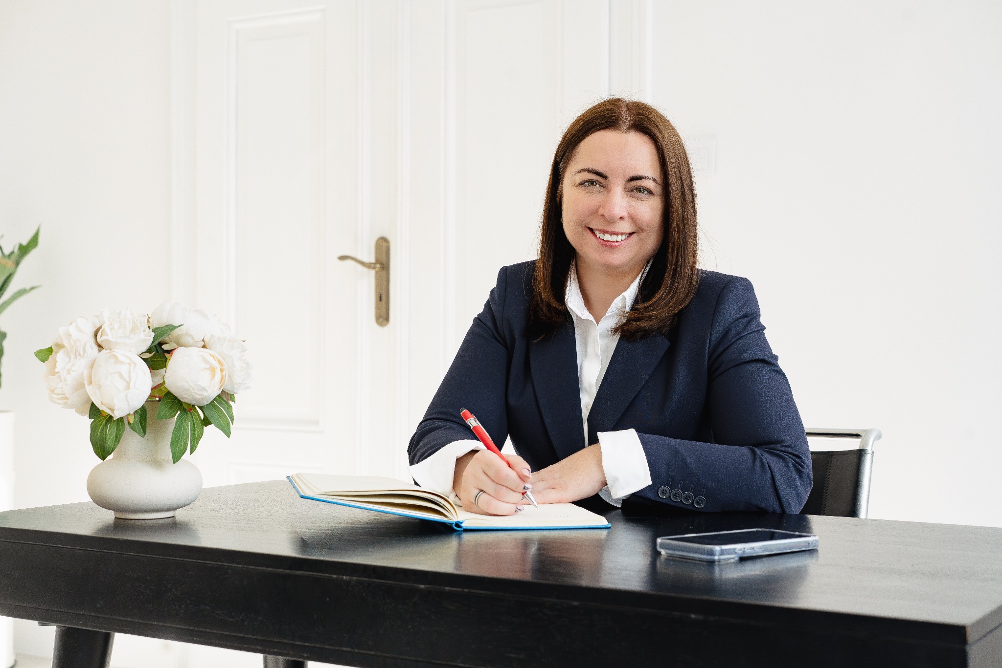 Uśmiechnięta kobieta w marynarce pisze w notatniku przy biurku z kwiatami i telefonem. Jasne wnętrze, profesjonalny wizerunek.