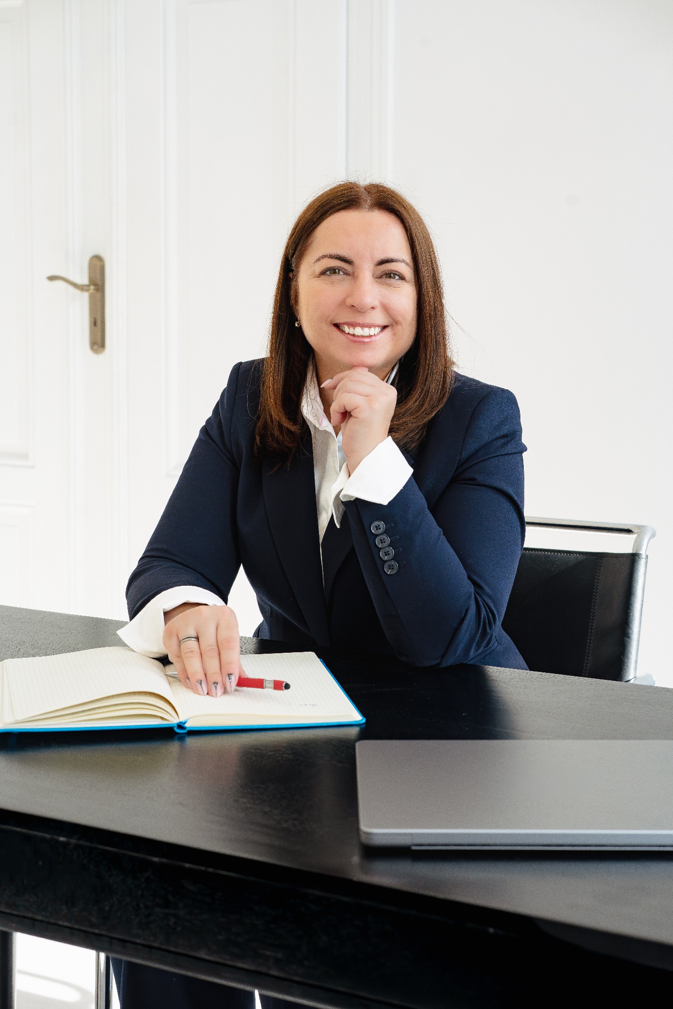 Uśmiechnięta kobieta w granatowym żakiecie siedzi za biurkiem z otwartym notatnikiem i czerwonym długopisem, opierając podbródek na dłoni. Widoczny laptop i białe drzwi.