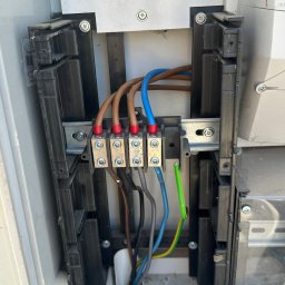 JB INSTALACJE ŁUKASZ BEDNAREK - Wnętrze skrzynki elektrycznej z podłączonymi przewodami: brązowe, niebieskie, zielono-żółte. Widoczne złącza śrubowe i elementy montażowe. Instalacja w szarej obudowie.