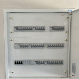 JB INSTALACJE ŁUKASZ BEDNAREK - Otwarta, biała skrzynka z bezpiecznikami Eaton na jasnej ścianie. Widoczne rzędy wyłączników. Drzwiczki skrzynki uchylone, ukazujące wnętrze.