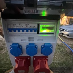JB INSTALACJE ŁUKASZ BEDNAREK - Skrzynka elektryczna z gniazdami siłowymi i licznikiem, zamontowana na zewnątrz, z widocznym budynkiem w tle i podłączonym przewodem.