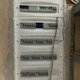 JB INSTALACJE ŁUKASZ BEDNAREK - Rozdzielnica elektryczna z zamontowanymi bezpiecznikami i przewodami, umieszczona na ścianie z surowym wykończeniem. Widoczne elementy instalacji i zabezpieczeń.
