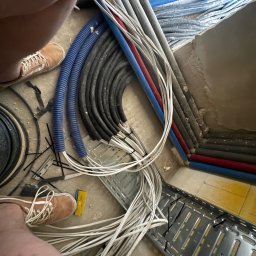 JB INSTALACJE ŁUKASZ BEDNAREK - Wiązka przewodów elektrycznych w peszlach i rurkach, ułożona w korycie kablowym. Widoczne fragmenty podłoża i butów osoby wykonującej prace.