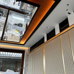 JB INSTALACJE ŁUKASZ BEDNAREK - Nowoczesne wykończenie wnętrza: podwieszany sufit z oświetleniem LED i dekoracyjna ściana z panelami. Widoczne elementy instalacji i okno z widokiem na zewnątrz.