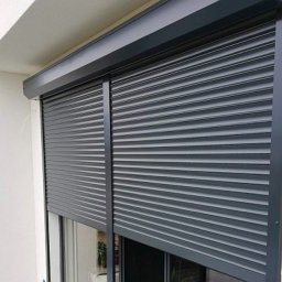 Oleh Bud - Grafitowe, częściowo opuszczone rolety zewnętrzne na oknie balkonowym, widoczne z zewnątrz, z delikatnym odbiciem otoczenia w szybie.