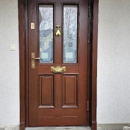 Oleh Bud - Eleganckie, brązowe drzwi wejściowe z szybami i złotymi detalami, takimi jak kołatka i szyld na list. Widoczne ościeżnice i fragment elewacji budynku.