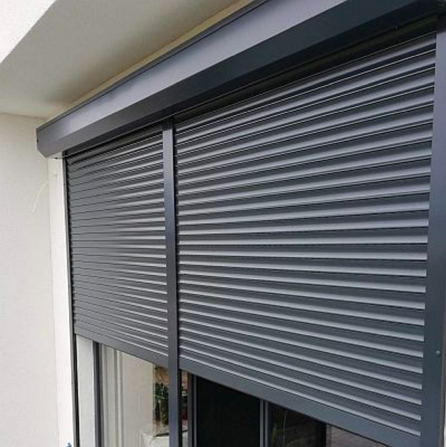 Grafitowe, częściowo opuszczone rolety zewnętrzne na oknie balkonowym, widoczne z zewnątrz, z delikatnym odbiciem otoczenia w szybie.