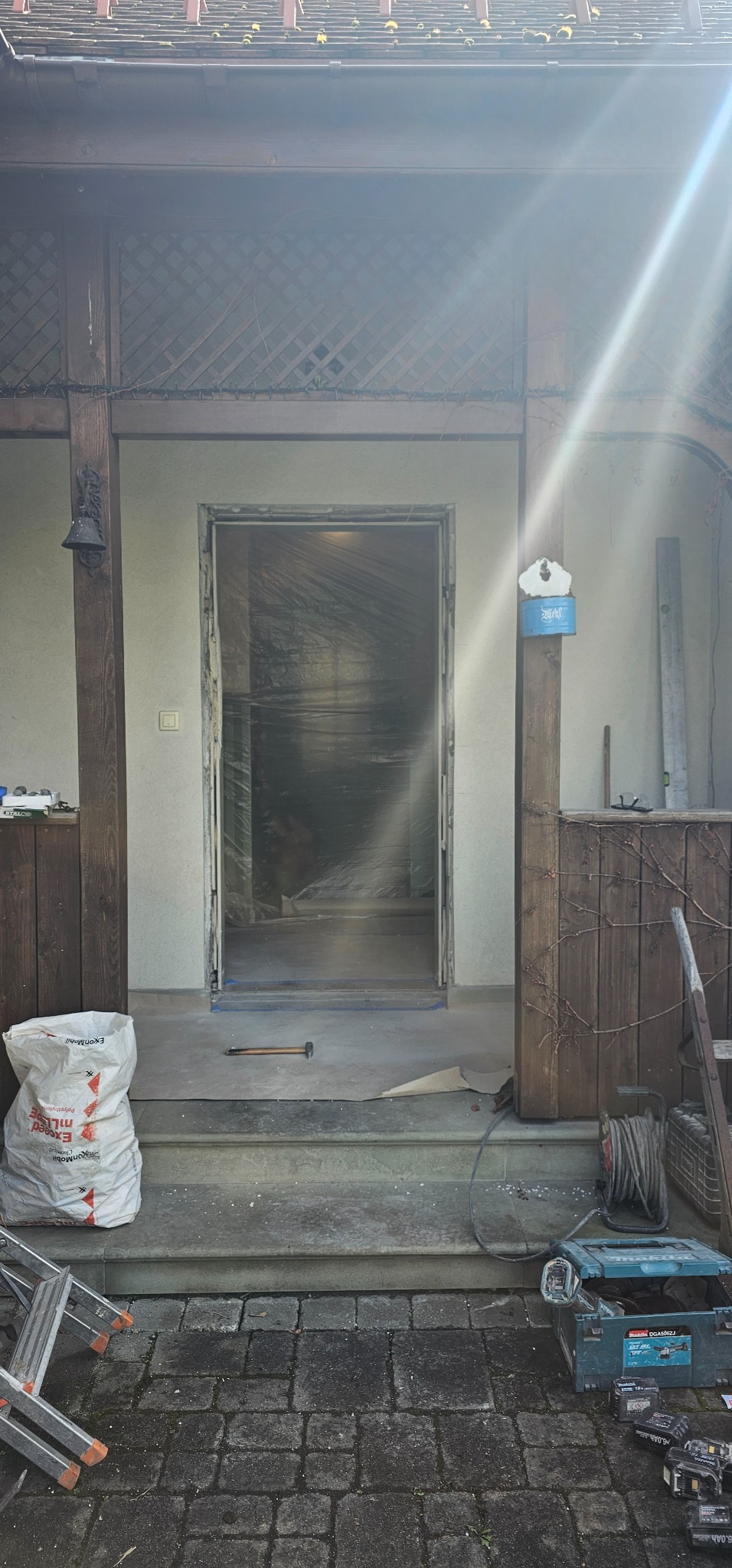 Przygotowanie do montażu drzwi wejściowych: widoczna surowa ościeżnica, narzędzia i materiały budowlane na schodach przed wejściem do domu.