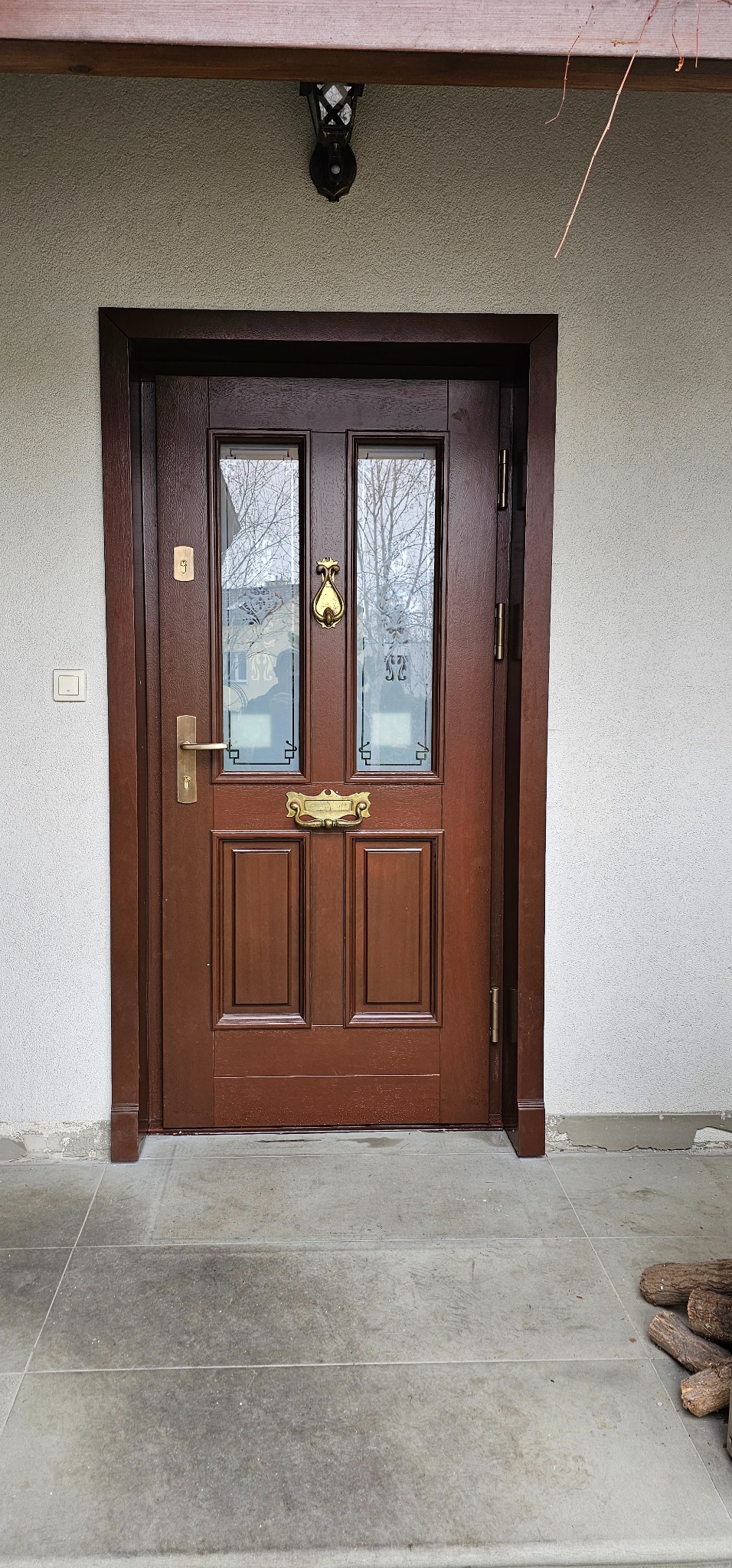 Eleganckie, brązowe drzwi wejściowe z szybami i złotymi detalami, takimi jak kołatka i szyld na list. Widoczne ościeżnice i fragment elewacji budynku.
