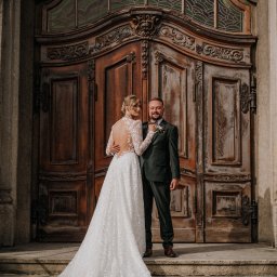 Miękka Sztuka Fotografia - Para młoda przed monumentalnymi, zdobionymi drzwiami z witrażem. Panna młoda w koronkowej sukni z odkrytymi plecami, pan młody w ciemnozielonym garniturze.