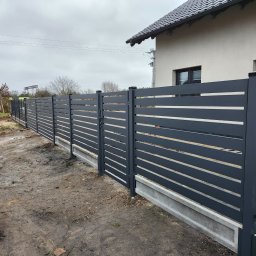 ogrodzenia pomorskie - Nowoczesne, poziome przęsła ogrodzeniowe w kolorze grafitowym, osadzone na betonowej podmurówce. W tle dom jednorodzinny i częściowo widoczny samochód dostawczy.
