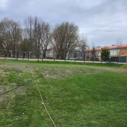 ogrodzenia pomorskie - Świeżo rozłożona trawa w rolkach na dużym terenie, widoczne linie łączeń i system nawadniania. W tle ogrodzenie panelowe i budynek.