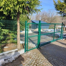 ogrodzenia pomorskie - Otwarta, zielona brama z siatki panelowej, zamontowana przy posesji z kostką brukową i zielonymi drzewami iglastymi. W tle widoczny budynek gospodarczy.