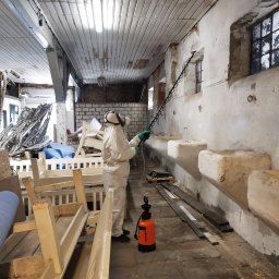Odgrzybianie-ozonowanie - Pracownik w kombinezonie ochronnym spryskuje ścianę wewnątrz budynku. Widoczny opryskiwacz i elementy wyposażenia wnętrza. Praca w toku.