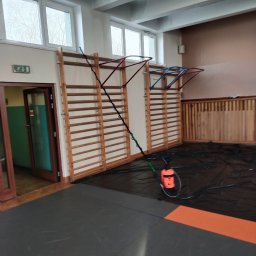 Odgrzybianie-ozonowanie - Pomarańczowy opryskiwacz ciśnieniowy z lancą na czarnej folii w sali gimnastycznej, przygotowany do zabiegu. Widoczne drabinki gimnastyczne i okna.