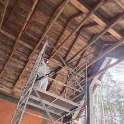 Odgrzybianie-ozonowanie - Pracownik w kombinezonie ochronnym na rusztowaniu, inspekcja drewnianej konstrukcji dachu w surowym stanie. Widok z dołu na drewniane belki i fragment lasu w tle.