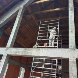 Odgrzybianie-ozonowanie - Pracownik w kombinezonie ochronnym na rusztowaniu, wykonujący prace na wysokości w surowym stanie budynku. Widoczne betonowe elementy konstrukcyjne i drewniany strop.