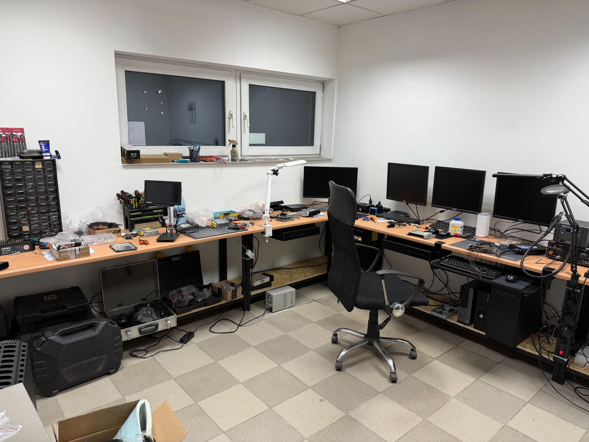 Warsztat elektronika z trzema monitorami, narzędziami i elementami elektronicznymi na biurku. Widoczne skrzynki narzędziowe i akcesoria. Jasne oświetlenie.