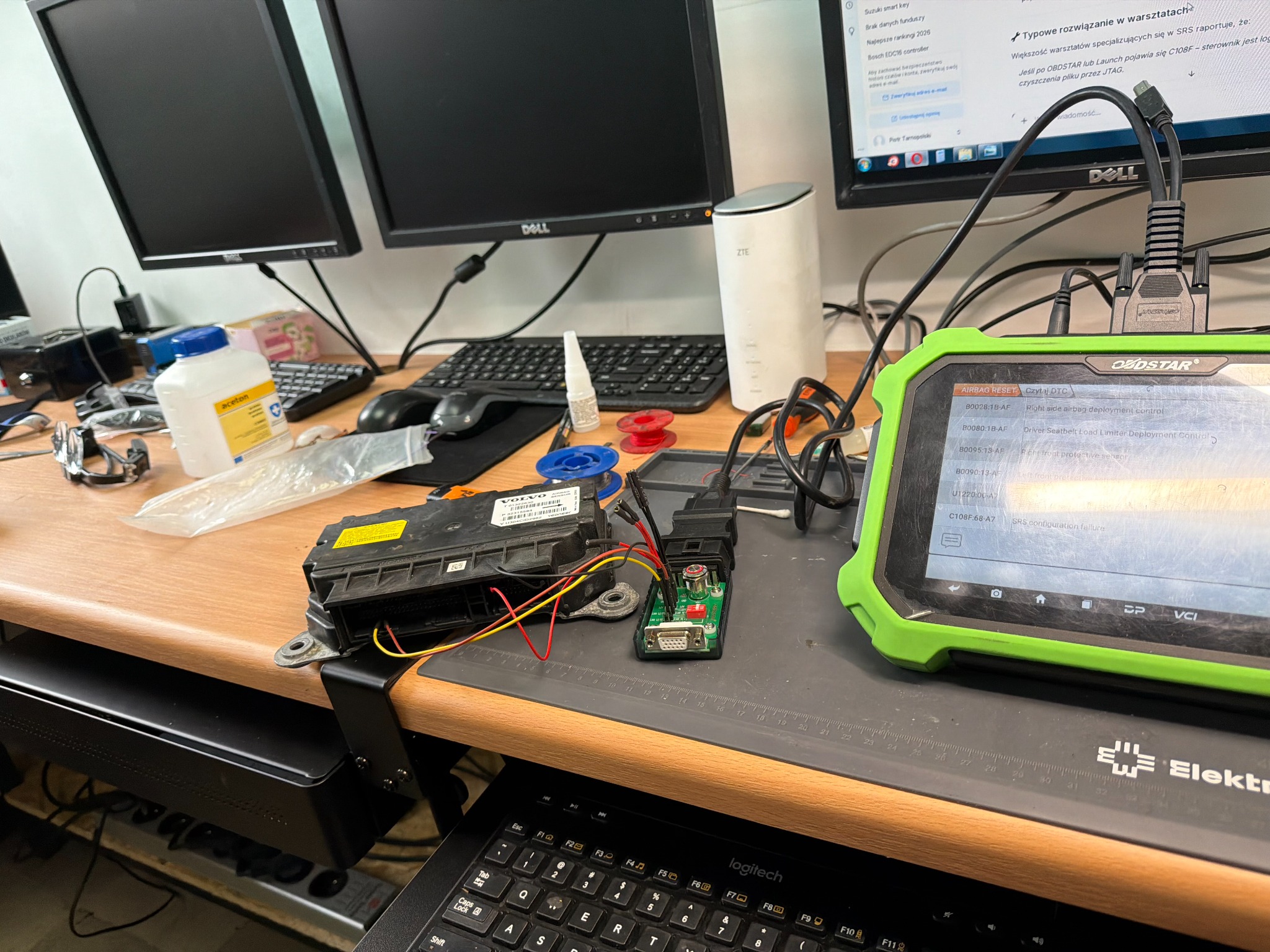 Diagnostyka modułu sterującego airbagiem za pomocą testera OBDSTAR. Widoczne kable, elektronika i monitory w tle. Warsztatowy stół z narzędziami.