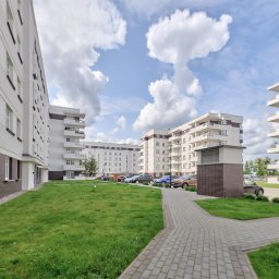 HOME MASTER NIERUCHOMOŚCI - Nowoczesne osiedle z białymi budynkami i balkonami, widok z poziomu trawnika. Na parkingu zaparkowane samochody, błękitne niebo z chmurami w tle.