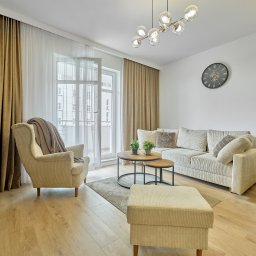 HOME MASTER NIERUCHOMOŚCI - Jasny salon z beżową sofą, fotelem i pufą. Okrągłe stoliki kawowe i dekoracyjny zegar na ścianie. Widok na balkon przez otwarte drzwi balkonowe.