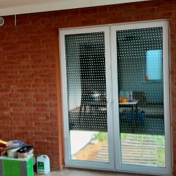 Tom-bud - Ściana z cegły klinkierowej w pokoju z białymi drzwiami balkonowymi z roletami. Widoczne materiały budowlane i narzędzia. Charakterystyczna faktura cegły.