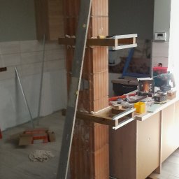 Tom-bud - Wnętrze w trakcie prac: kolumna z cegły z drewnianymi elementami, metalowa listwa oparta o nią, narzędzia i materiały budowlane na blacie. Etap wykończenia.