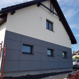 Art Bud - Dwukolorowa elewacja domu z szarymi panelami na dole i jasną górą. Widoczne rusztowania i okna. Dach czarny, spadzisty. Błękitne niebo w tle.