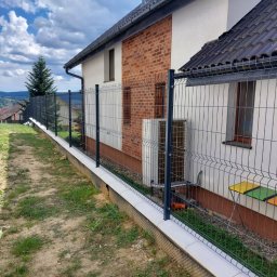 Krystian Cebula - Panelowe ogrodzenie z podmurówką na nierównym terenie, widok z boku. W tle dom z ceglaną elewacją i zielone wzgórza. Słoneczny dzień, częściowe zachmurzenie.