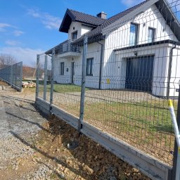 Krystian Cebula - Nowoczesne ogrodzenie panelowe z podmurówką betonową, szara siatka, widok na biały dom z garażem i balkonem, częściowo zagospodarowany teren.