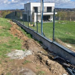Krystian Cebula - Nowoczesne ogrodzenie panelowe z podmurówką w Nowym Sączu, widok z ziemi, na tle domu i błękitnego nieba z chmurami. Widoczne elementy montażowe.