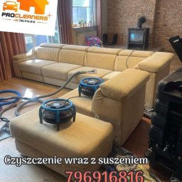 PROCLEANERS - Beżowa, narożna kanapa po czyszczeniu, z dwoma wentylatorami na siedzisku. W tle ceglana ściana i kominek. Widoczne węże i urządzenie czyszczące.