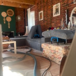 PROCLEANERS - Pranie tapicerki kanapy w loftowym wnętrzu. Widoczny sprzęt do prania i suszenia tapicerki. W tle ceglana ściana z roślinami i dekoracjami.