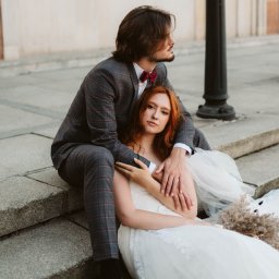 Krzysztof Pszczółkowski - Para młoda siedzi na schodach: on w kraciastej marynarce i bordowej muszce, ona w białej sukni ślubnej z bukietem. Romantyczna sesja w miejskiej scenerii.