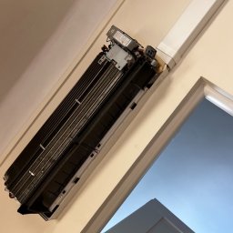 Freezzzler Barański J,. Cymerman K., Nalazek K. Spółka Jawna - Otwarty klimatyzator ścienny z widocznymi elementami wewnętrznymi, zamontowany nad drzwiami w jasnym pomieszczeniu. Widoczne przewody i elementy instalacji.