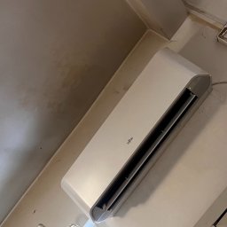 Freezzzler Barański J,. Cymerman K., Nalazek K. Spółka Jawna - Biały klimatyzator Fujitsu zamontowany na ścianie, widoczny kabel zasilający i fragment kratki wentylacyjnej. Ujęcie z dołu, lekko zniekształcona perspektywa.