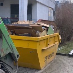 Fajans24 kontenery l wrocław - Żółty kontener, wypełniony odpadami poremontowymi (drewno, płyty, kartony), z numerem 791 991 650 na boku. Obok częściowo widoczne zielone ramię załadunkowe.